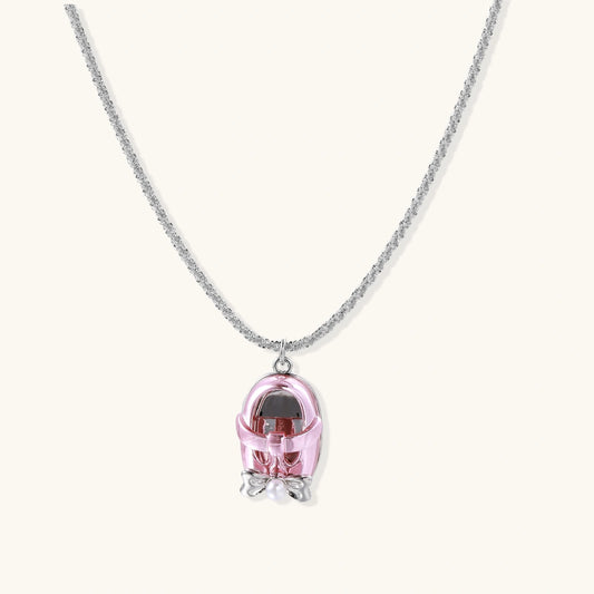 Pink baby-shoe pendant necklace with a silver-tone bow and pearl accent on a pale background.